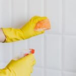 A person wearing yellow gloves cleans bathroom tiles with a sponge and spray bottle.